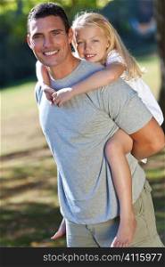 A young father with his blond daughter on his shoulders having fun in a sun bathed green park