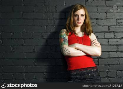 A young female with full arm tattoo leaning up against a black brick wall.