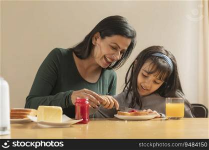 A YOUNG GIRL CHEERFULLY SPREADING JAM ON A SLICE OF BREAD WITH MOTHER SITTING BESIDE