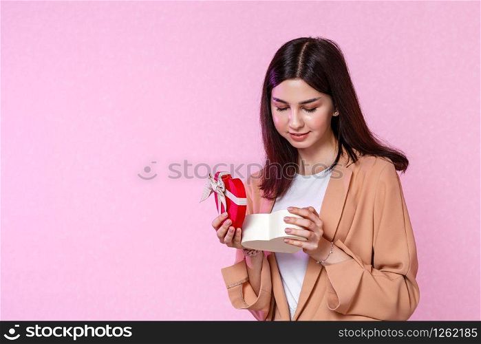 a young girl in a beige jacket with a gift box in her hands. surprised and happy with the gift. on a pink isolated background.