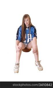 A young girl in a blue football outfit sitting on her football for a rest, withher long brunette hair and sneakers, for white background.