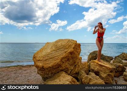 a young girl in a closed red swimsuit with a retro camera and an exposure meter is taking pictures while standing on a rocky beach