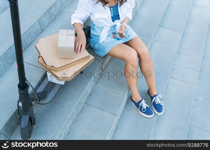 A young girl is sitting on the stairs near her scooter and a lot of boxes she took from the post office. The concept of delivery, ordering from online stores. A young girl is sitting on the stairs near her scooter and a lot of boxes she took from the post office. The concept of delivery, ordering from online stores.