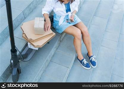 A young girl is sitting on the stairs near her scooter and a lot of boxes she took from the post office. The concept of delivery, ordering from online stores. A young girl is sitting on the stairs near her scooter and a lot of boxes she took from the post office. The concept of delivery, ordering from online stores.