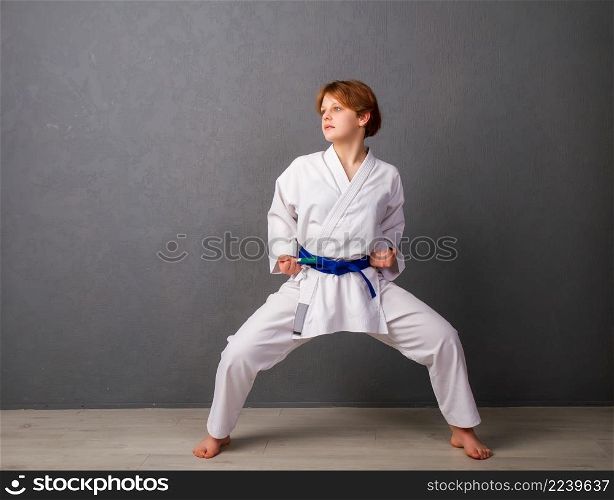 A young girl karateka in a white kimono and a blue belt trains and performs a set of exercises against a gray wall