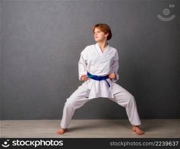 A young girl karateka in a white kimono and a blue belt trains and performs a set of exercises against a gray wall