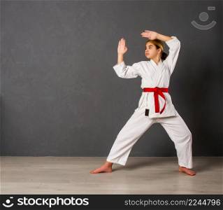 A young girl karateka in a white kimono and a red belt trains and performs a set of exercises against a gray wall