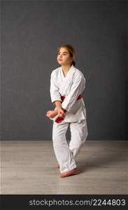 A young girl karateka in a white kimono and a red belt trains and performs a set of exercises against a gray wall