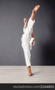 A young girl karateka in a white kimono and a red belt trains and performs a set of exercises against a gray wall