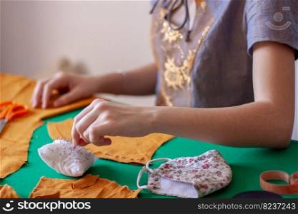 A young girl seamstress takes a needle from a pillow for needles. Orange cloth on a green table. Focus on a pad with needles and fingers. Nearby lies a face mask. Horizontal.