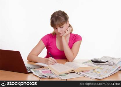 A young girl sits at a table with a pile of newspapers, encyclopedias and a laptop