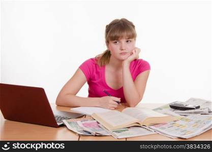 A young girl sits at a table with a pile of newspapers, encyclopedias and a laptop