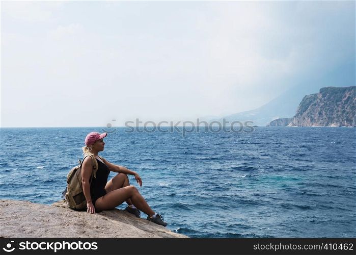 A young girl with blond hair sits on top of a mountain and looks at the sea and beach. Relax and travel on nature.. A young girl with blond hair sits on top of a mountain and looks