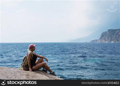 A young girl with blond hair sits on top of a mountain and looks at the sea and beach. Relax and travel on nature.. A young girl with blond hair sits on top of a mountain and looks