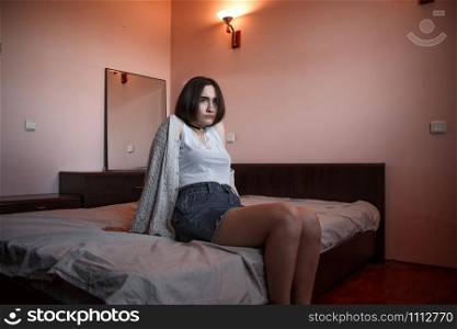 a young girl with short hair in the sweater and denim skirt sitting alone on the bed in the bedroom in a depression