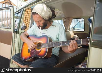 A young hippie plays the guitar while smoking a cigarette while he sits in the side entrance of his old classic travel van.