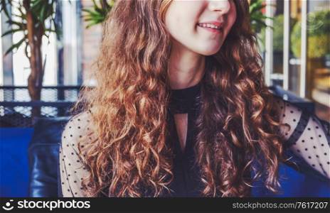 A young lady with curly brown hair rests in the cafeteria, leaning on the back of the chair and looking away. We only see the lower half of her face that looks away from the camera with a light smile on her lips. Toned image.. A young lady with curly brown hair rests in the cafeteria, leaning on the back of the chair and looking away from the camera with a smile.