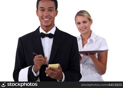 a young latino well dressed waiter writing order and a young blonde waitress holding a platter