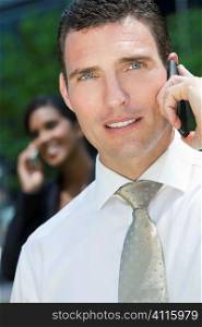 A young male executive talking on his cell phone with his female colleague on her phone out of focus in the background