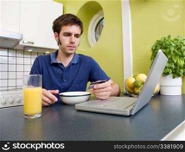 A young male shops online with a credit card in his kitchen while eating breakfast