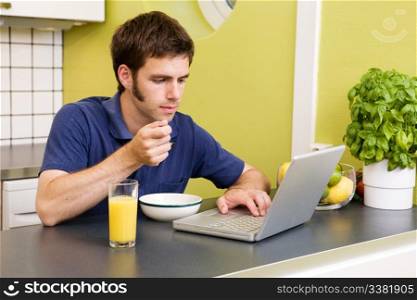A young male works on the computer in the kicthen while eating breakfast
