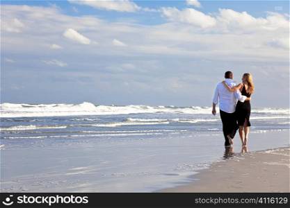 A young man and woman arms around each other walking as a romantic couple on a beach
