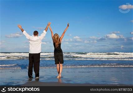 A young man and woman celebrating arms raised and holding hands as a romantic couple on a beach with a bright blue sky