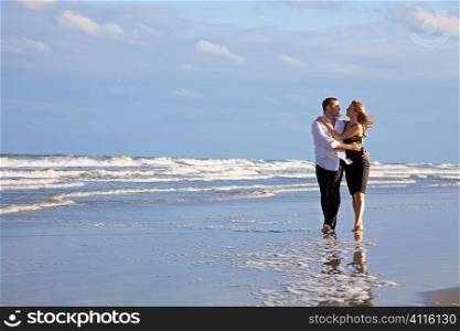 A young man and woman couple having romantic fun walking and embracing on a beach with a bright blue sky