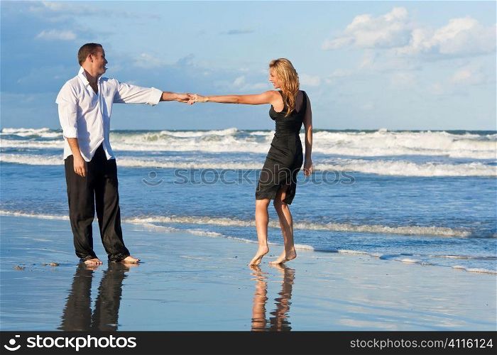 A young man and woman holding hands and having fun dancing as a romantic couple on a beach with a bright blue sky and sea