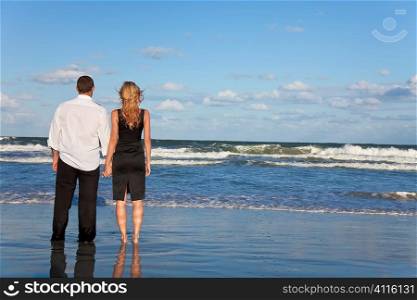 A young man and woman holding hands as a romantic couple looking out to sea on a beach with a bright blue sky