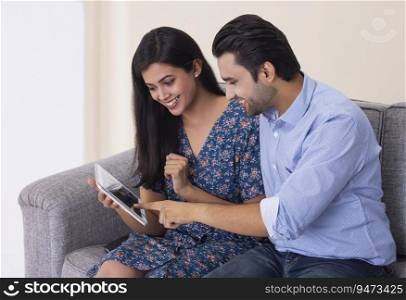 A young man and woman sitting together in their room selecting dresses on a tablet phone.