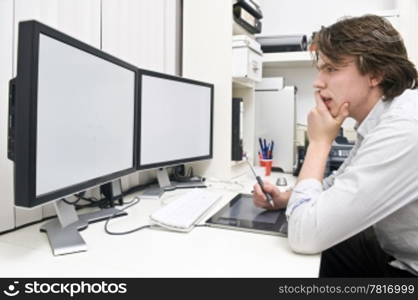 A young man at work behind a dual monitor work station in a design studio office environment