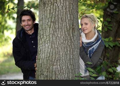 a young man dressed in coat and a young blonde woman posing near a tree