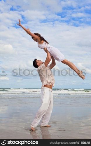 A young man holding his girlfriend above is head as she jumps in celebration on a beach