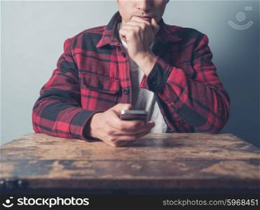 A young man in a checkered jacket is sitting at a desk and is using a smartphone