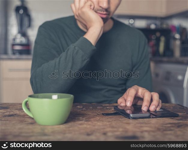 A young man is drinking coffee and using a smart phone in a kitchen