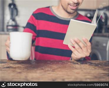 A young man is reading a book and drinking coffee in a kitchen