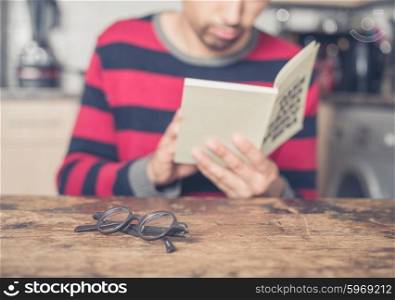 A young man is reading a book in a kitchen. Focus on his reading glasses on the table in front of him.