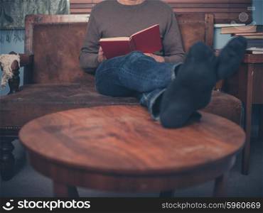 A young man is relaxing on a sofa at home and is reading a book