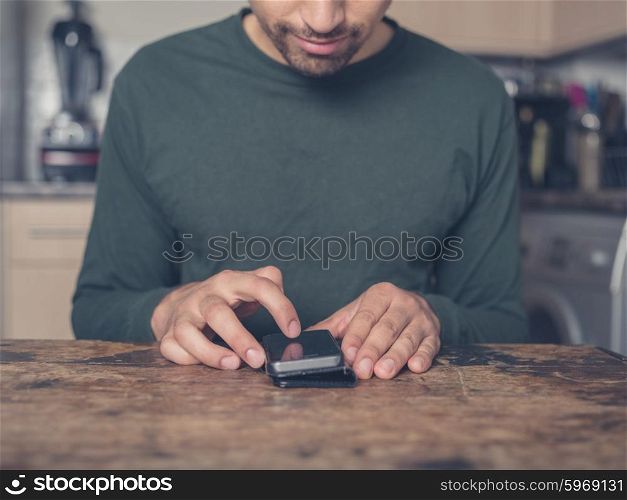 A young man is sitting at a table in a kitchen and is using a smart phone