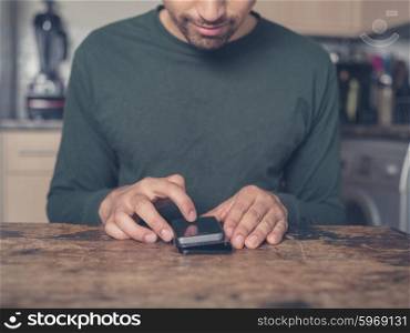 A young man is sitting at a table in a kitchen and is using a smart phone