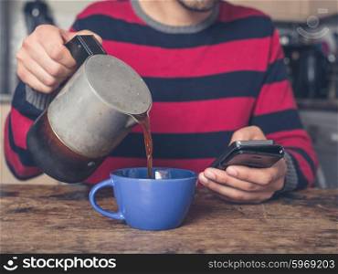 A young man is sitting at a table in a kitchen and is pouring coffee while using a smart phone