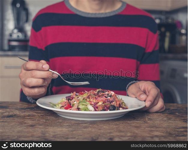 A young man is sitting at a table in a kitchen and is eating salad