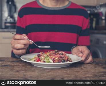 A young man is sitting at a table in a kitchen and is eating salad