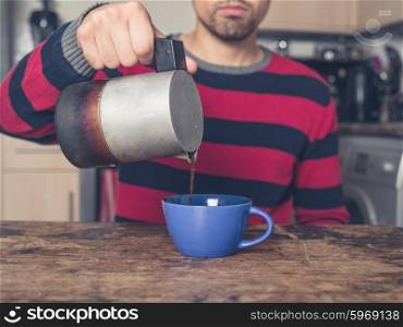A young man is sitting at a table in a kitchen and is pouring coffee