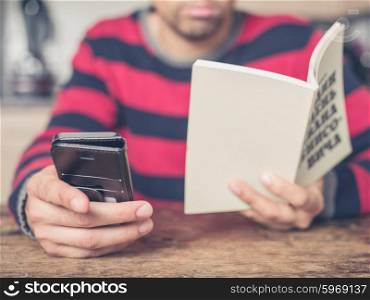 A young man is sitting at a table in a kitchen reading a book and using a smart phone