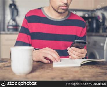 A young man is sitting at a table in a kitchen reading a book and using a smart phone