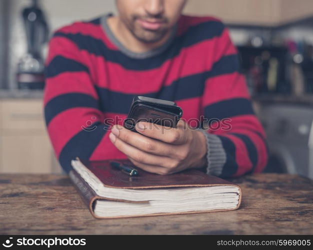 A young man is sitting at a table in a kitchen with a notebook and is using a smart phone