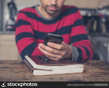 A young man is sitting at a table in a kitchen with a notebook and is using a smart phone