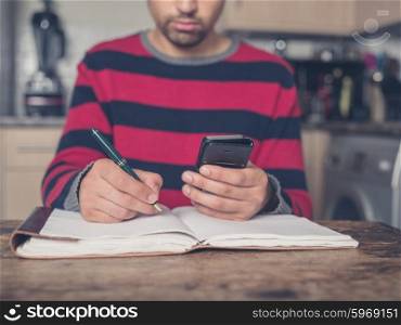 A young man is sitting at a table in a kitchen with a notebook and is using a smart phone